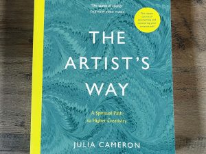 Text displays a copy of the book, The Artist's Way: A Spiritual to Higher Creativity resting on a wooden background.